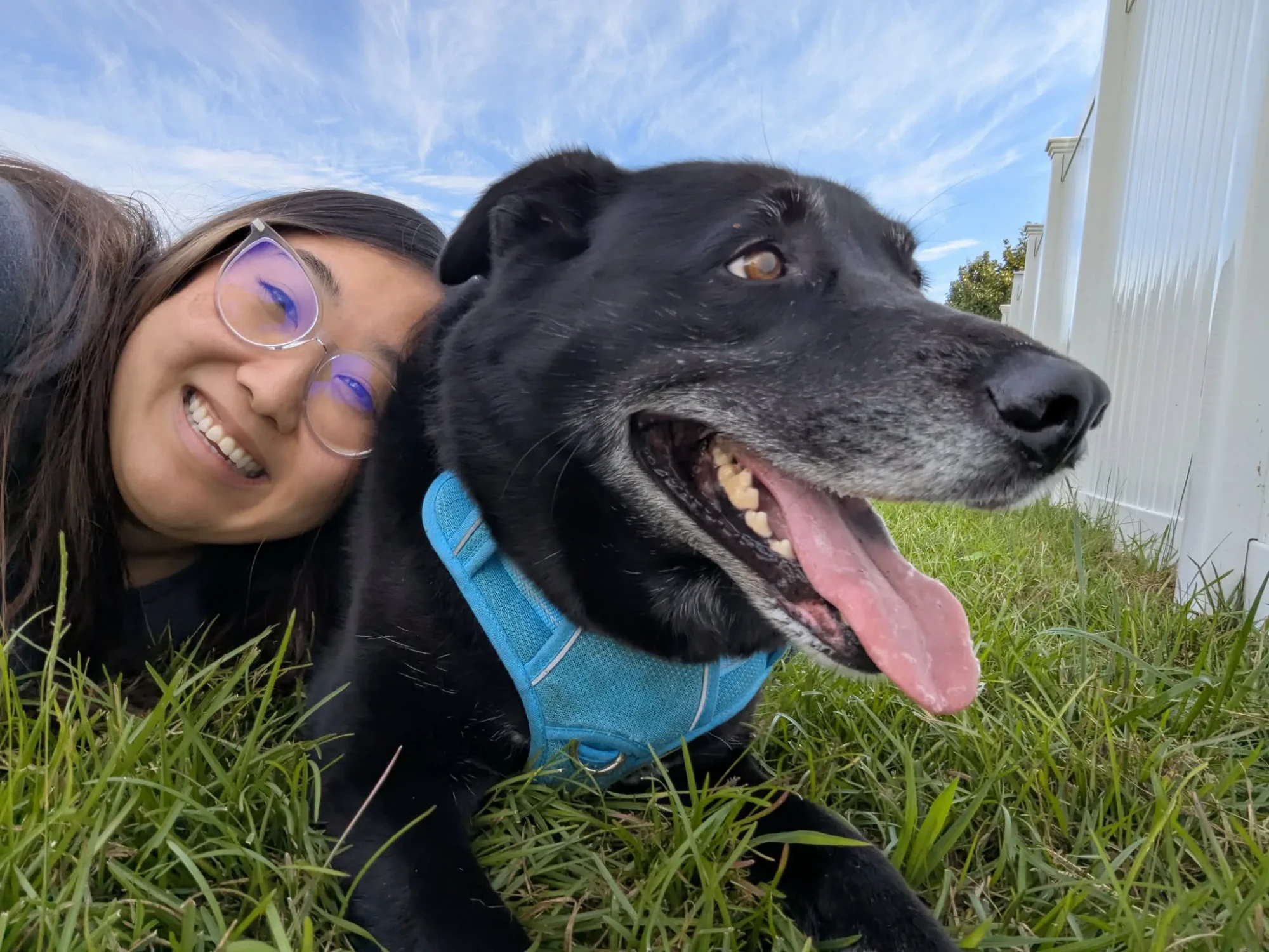 Wan Dee lying in the grass next to her black dog. The dog is grinning with her tongue out.